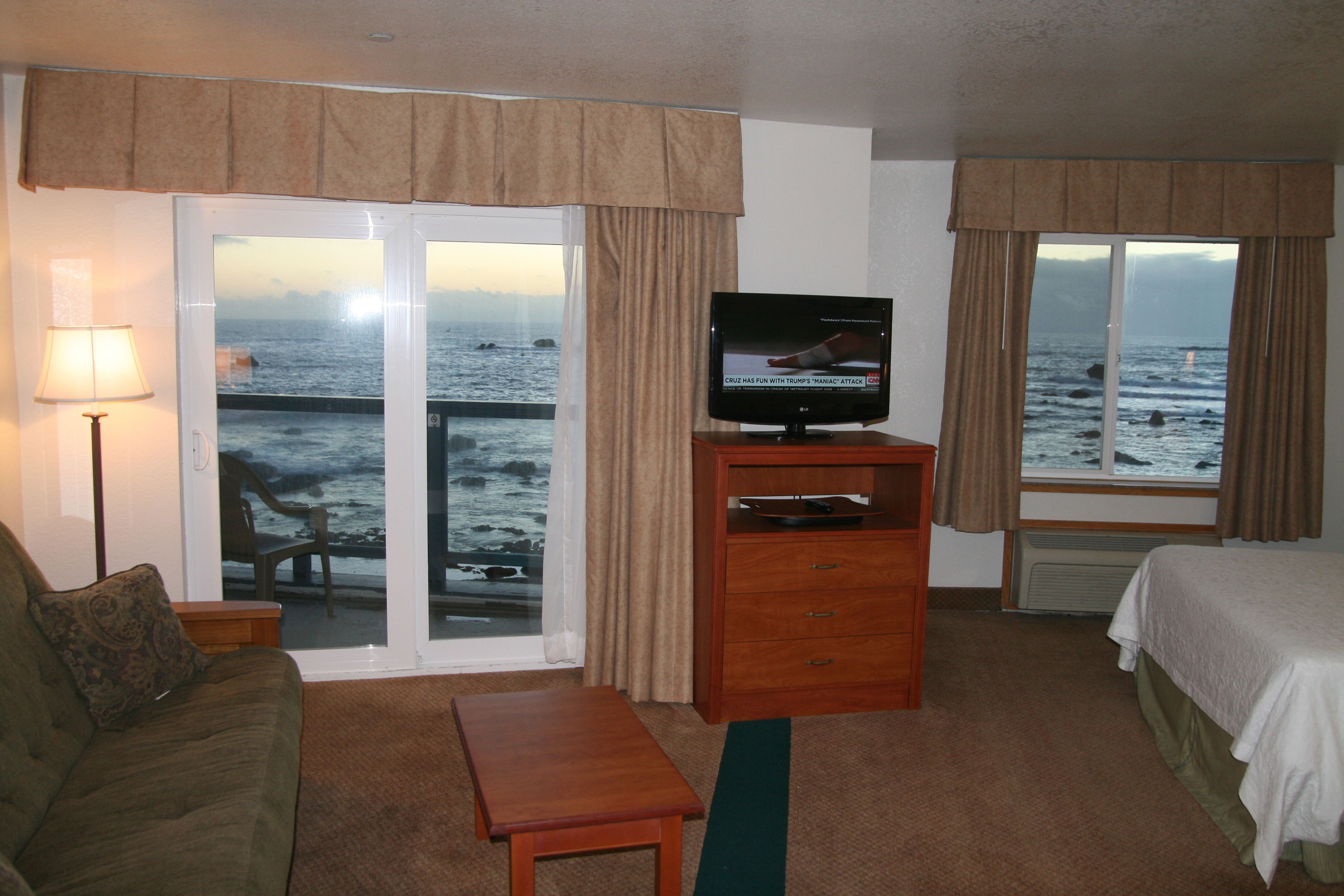 A hotel room with a bed, a television on a wooden stand, a sofa, a small table, and a view of the ocean through a sliding glass door.