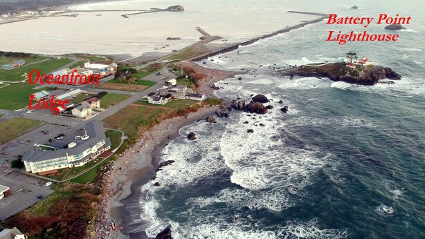 An aerial view of Battery Point Lighthouse and Oceanfront Lodge, featuring the lighthouse on a rocky outcrop and the lodge with a curved roof and adjacent parking area.