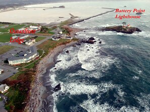 Vista aérea del faro de Battery Point y el Oceanfront Lodge, mostrando el faro sobre un promontorio rocoso y el lodge con techo curvo y zona de estacionamiento contigua.