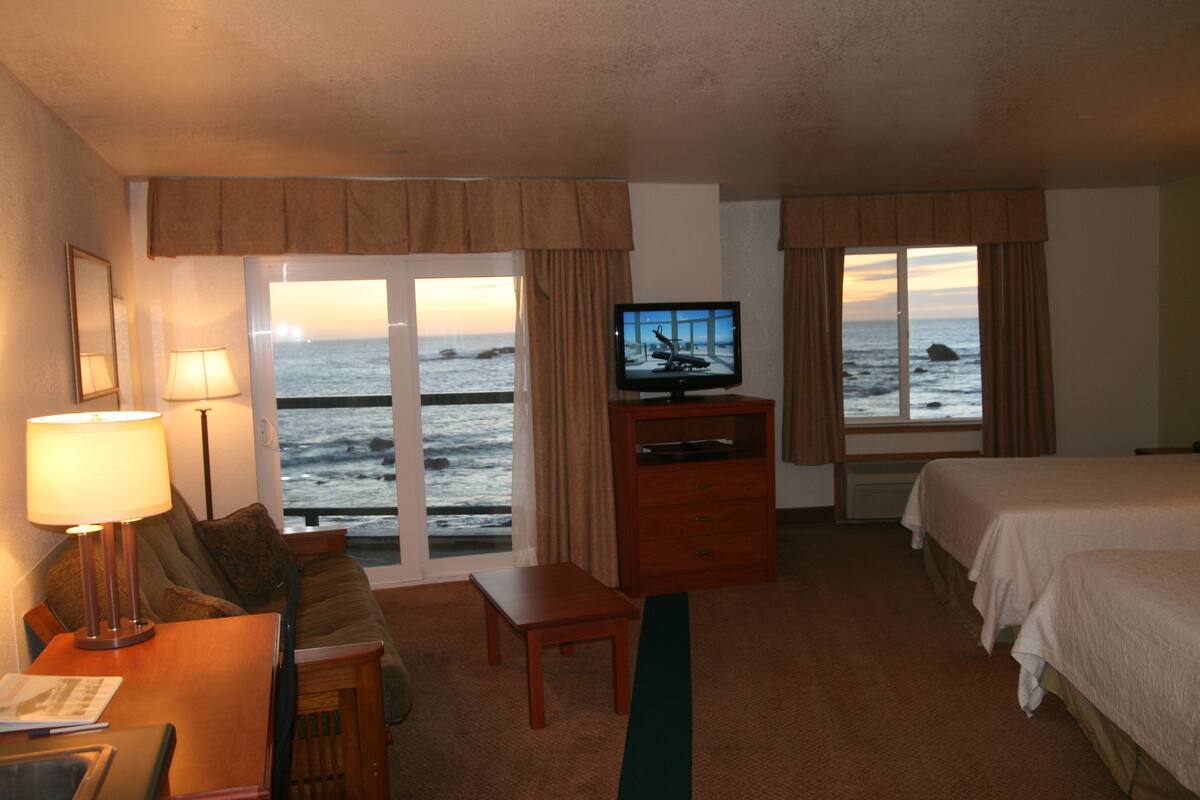 A hotel room with a bed, a desk with a lamp, a television, and a view of the ocean.