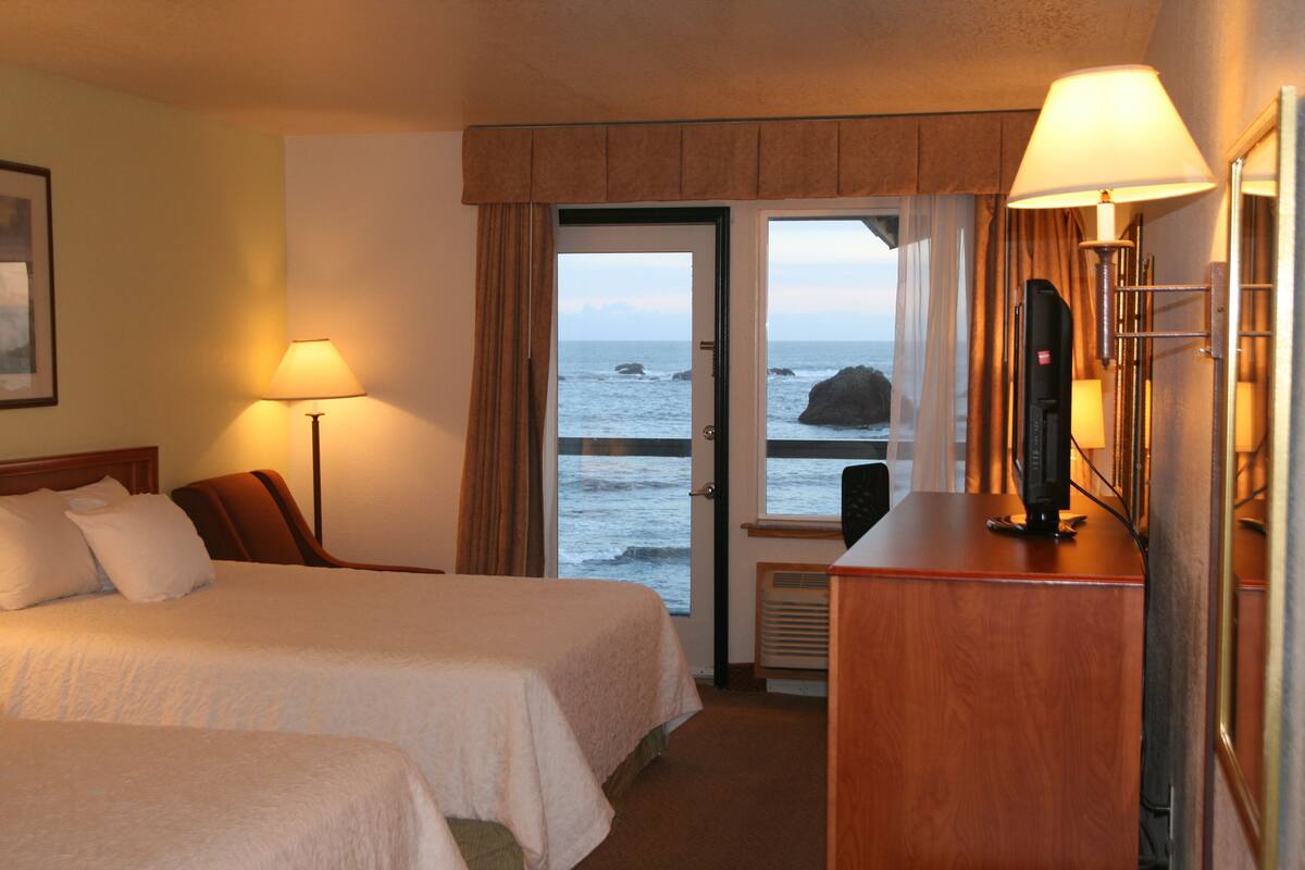 A hotel room with two beds, a desk with a computer, a lamp, and a view of the ocean through a door.