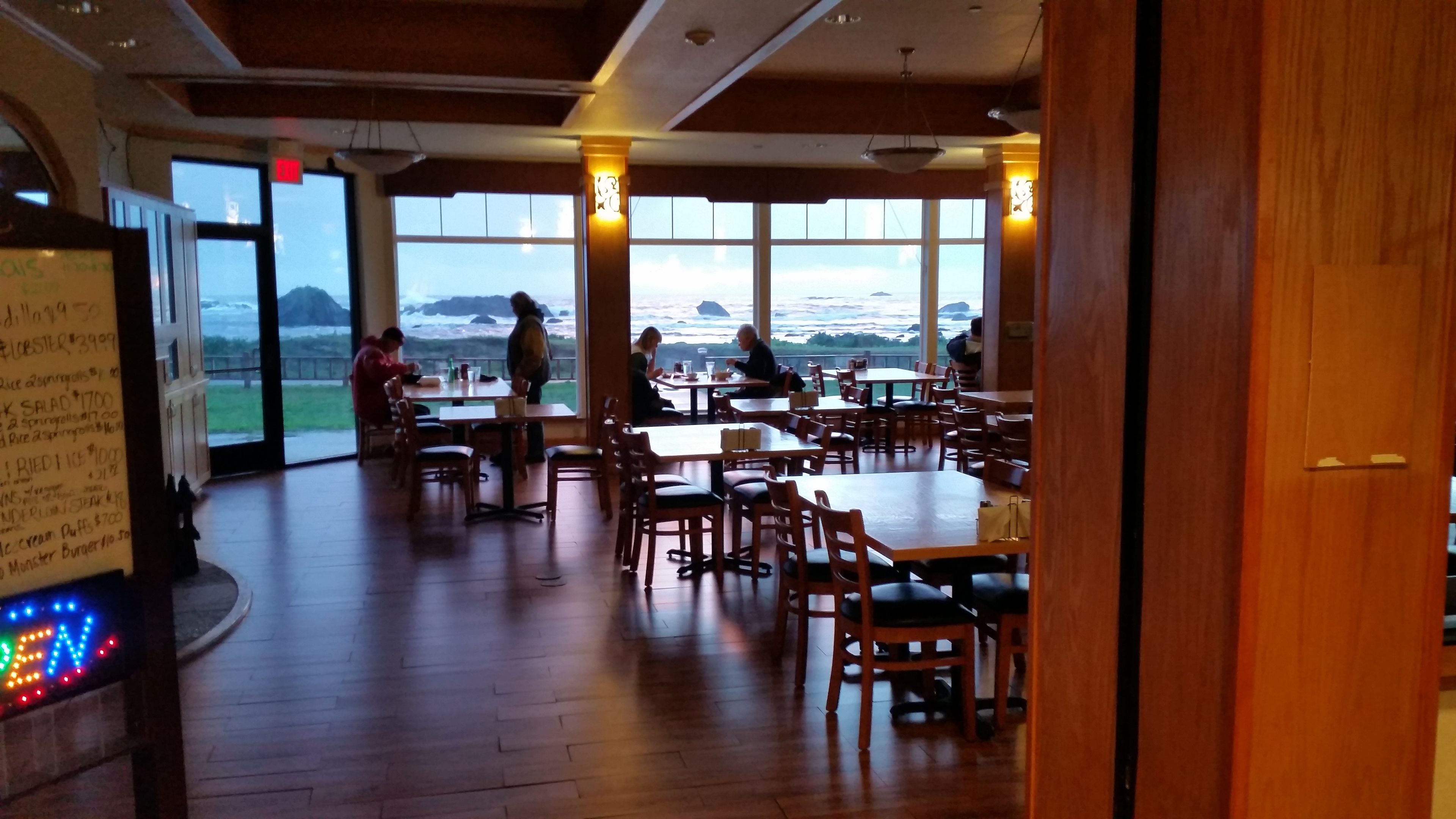 Een restaurant met houten vloeren en grote ramen met uitzicht op zee. Tafels en stoelen staan klaar voor diners.