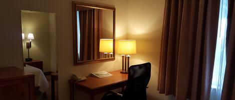 Habitación de hotel con escritorio, dos lámparas, teléfono, silla, espejo y ventana con cortinas.