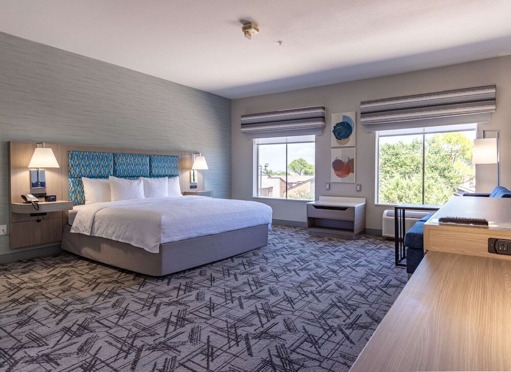Una habitación de hotel moderna con una cama grande, mesitas de noche con lámparas, un escritorio y vistas al exterior a través de amplias ventanas.