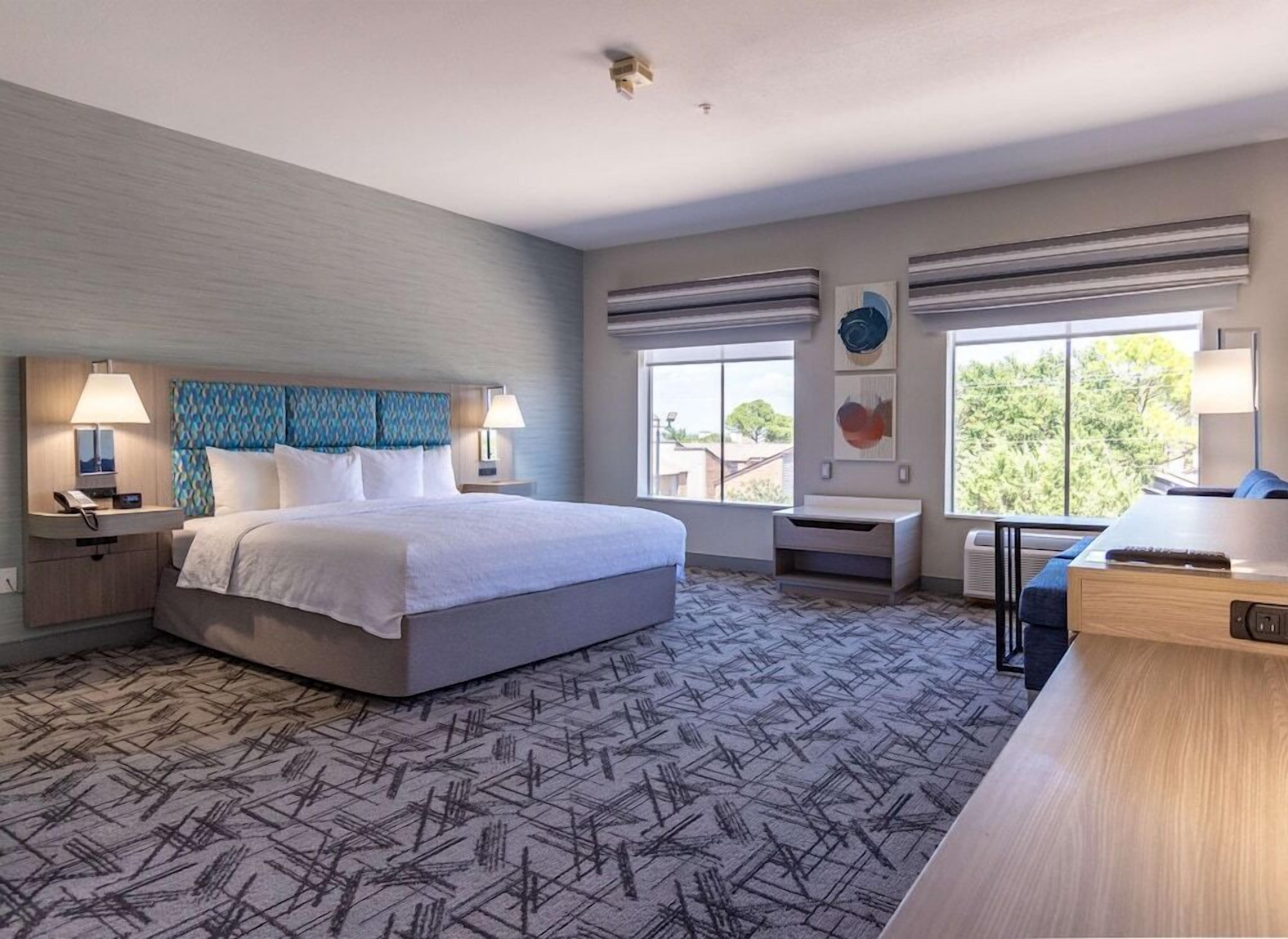 A modern hotel room with a large bed, bedside tables with lamps, a desk, and a view of the outdoors through large windows.