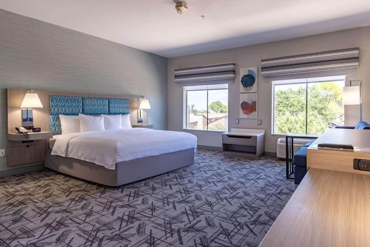 A hotel room with a large bed, bedside tables with lamps, a desk, and a view of the outdoors through large windows.