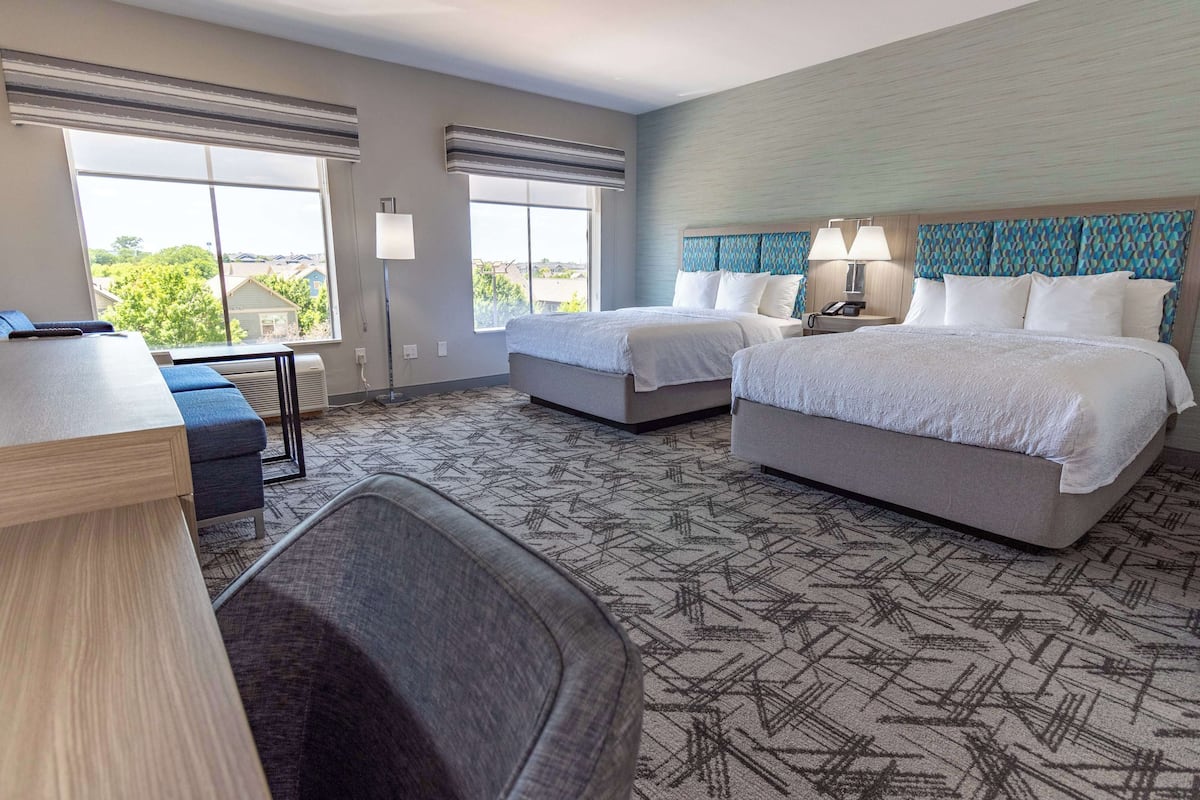 A hotel room with two beds, a desk, and a chair. There are large windows and a view of the outdoors.