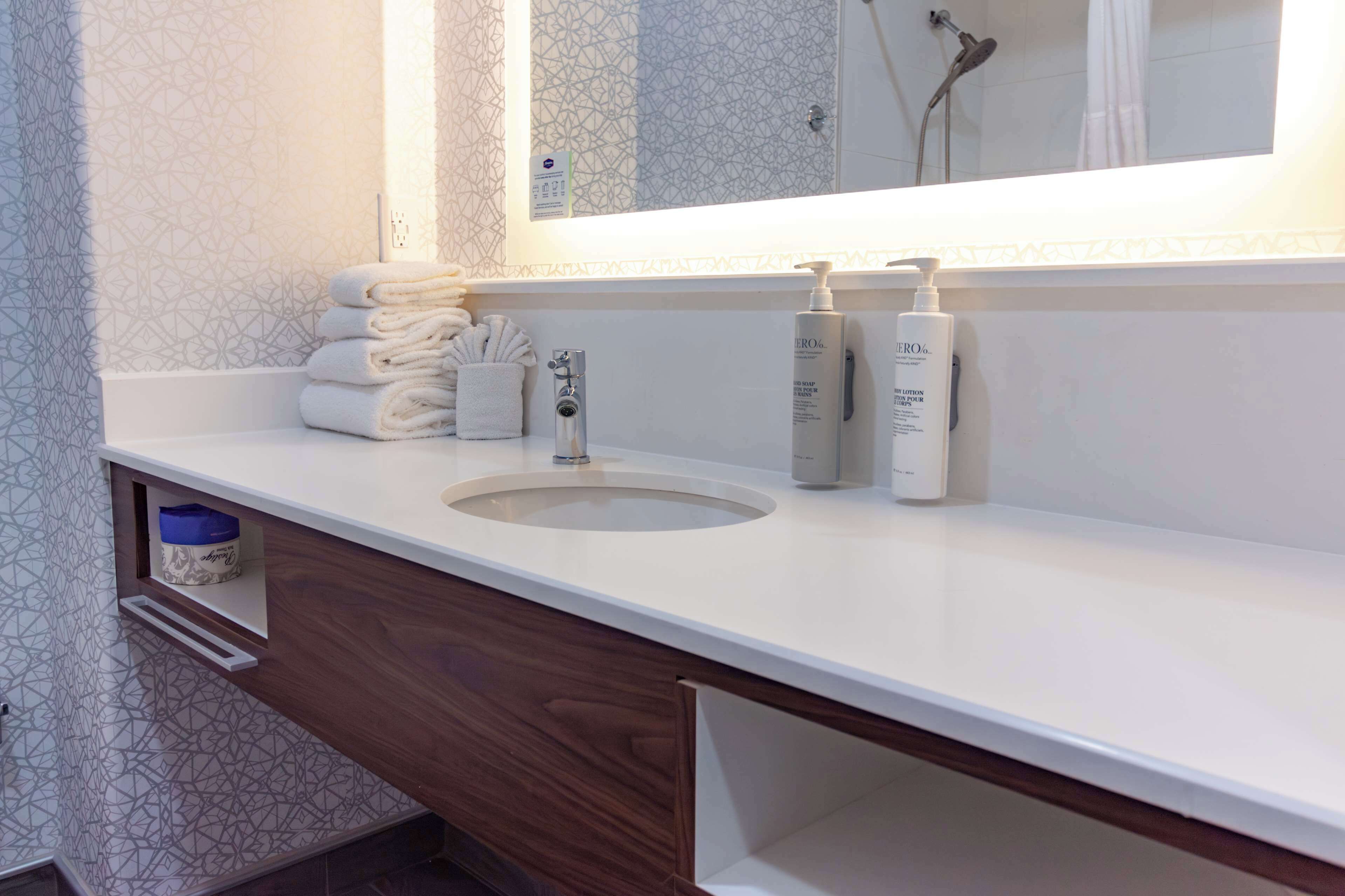 Une salle de bain avec un lavabo blanc, un miroir et un porte-serviettes. On peut voir deux flacons de lotion sur le comptoir.