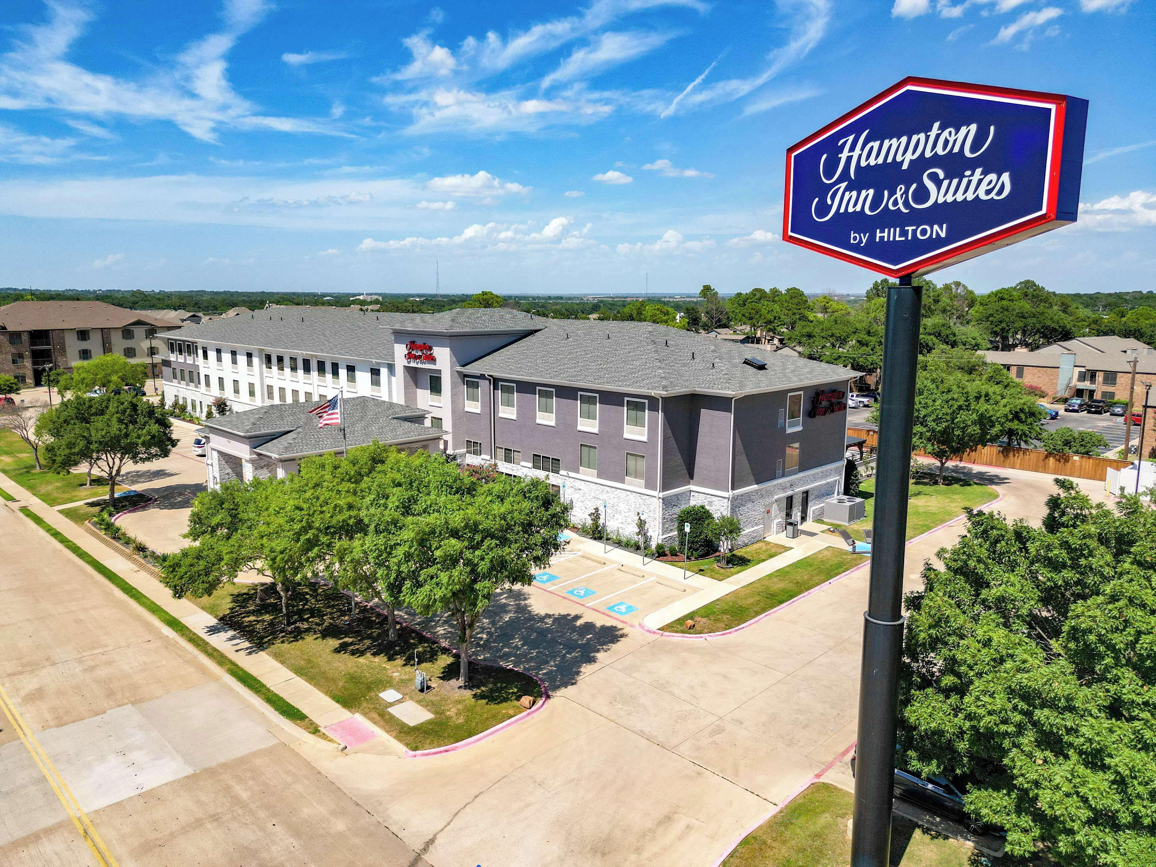 A Hampton Inn & Suites by Hilton building with a parking lot, flag, and surrounding greenery.