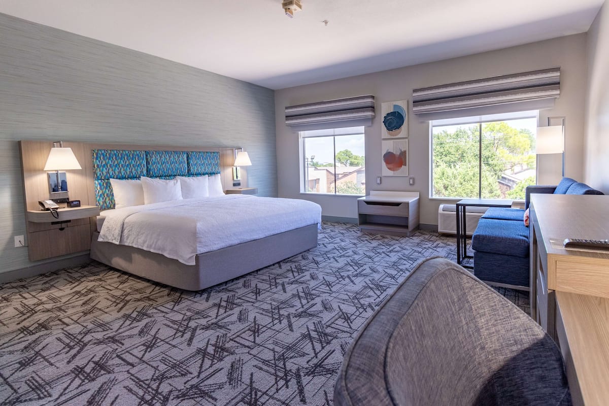 A hotel room with a large bed, two bedside tables with lamps, a desk with a chair, and a view of the outdoors through large windows.