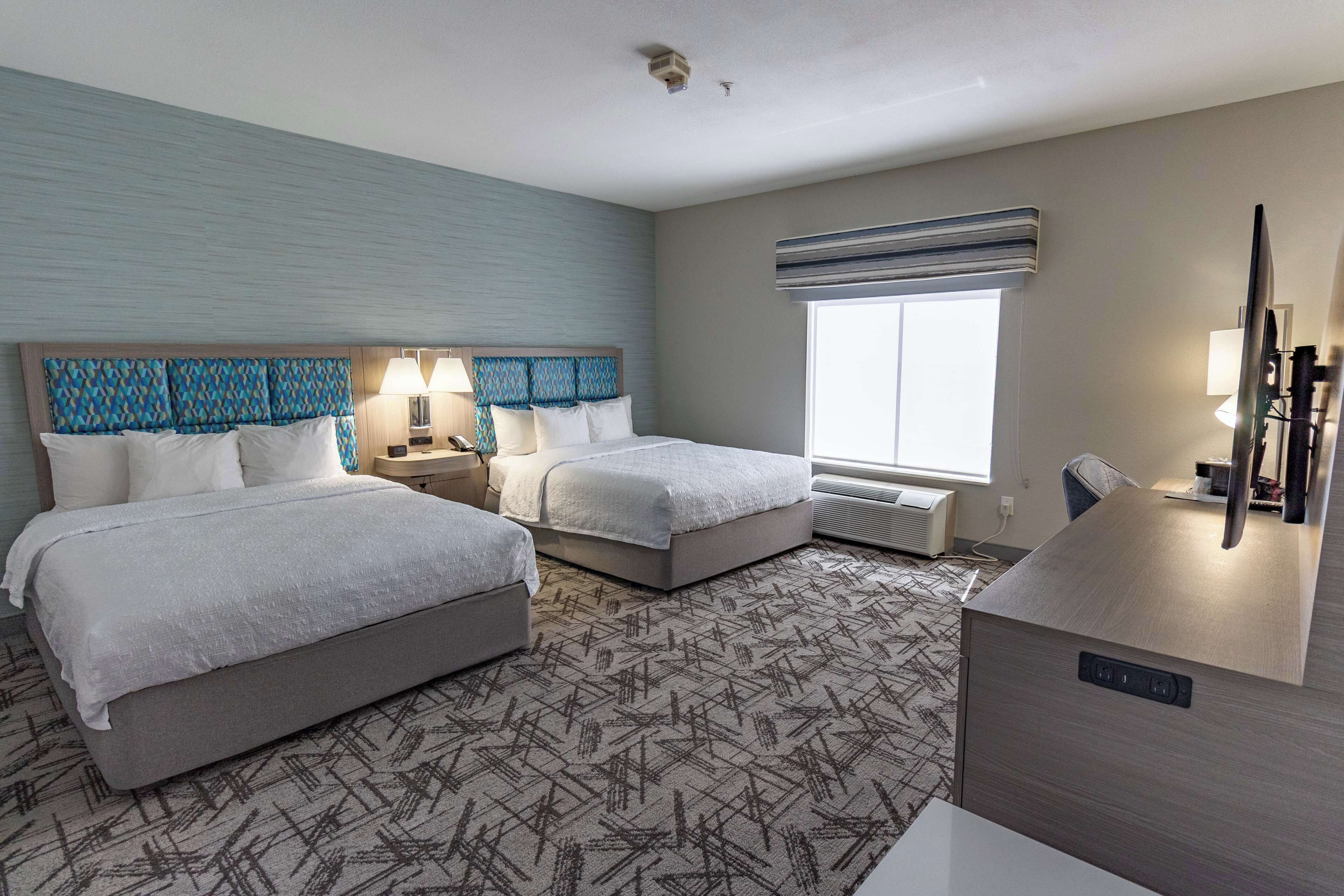 A hotel room with two beds, a desk, a chair, a lamp, and a large window.