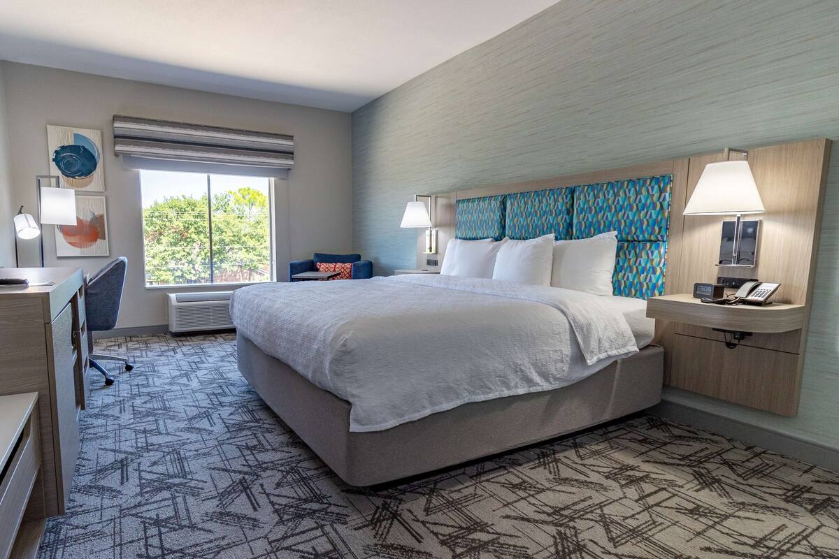 A hotel room with a large bed, a desk, a chair, a window with a view of trees, and a patterned carpet.