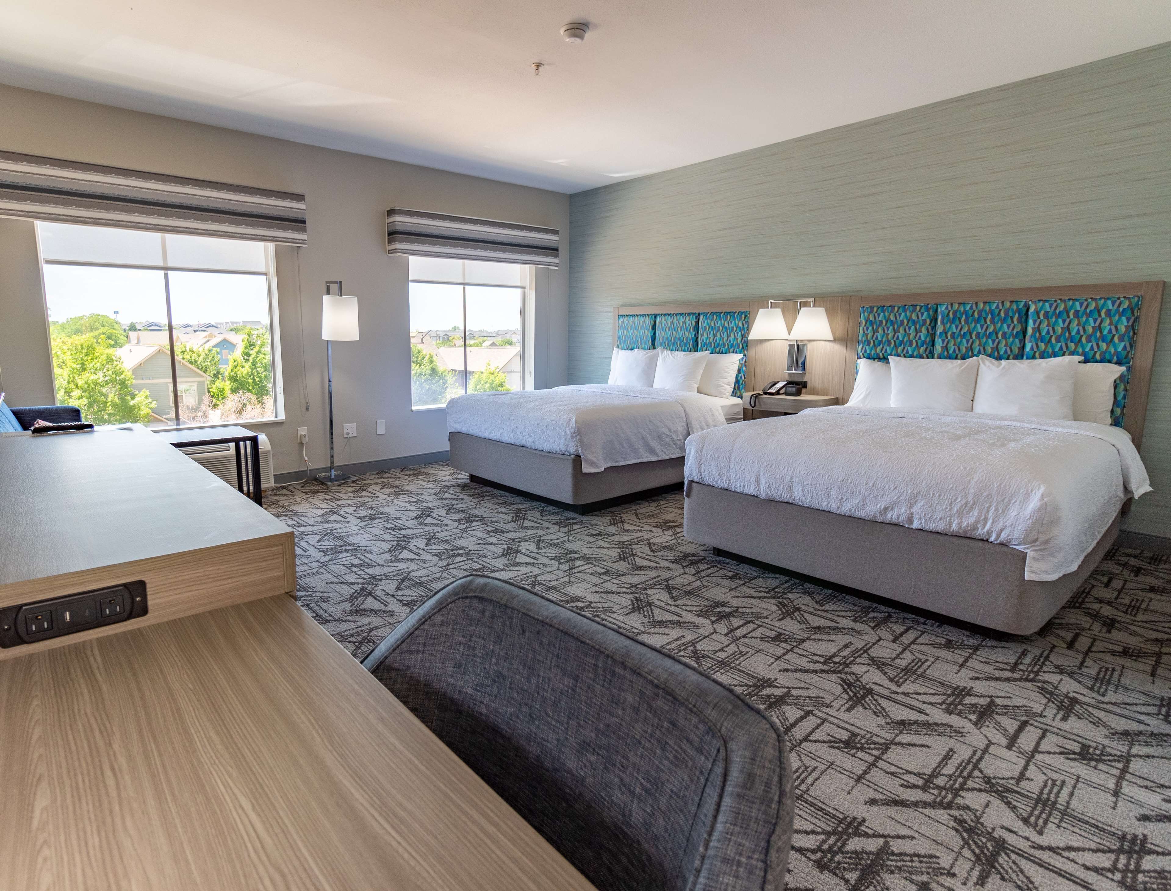 A hotel room with two beds, a desk, and a view of the outside.
