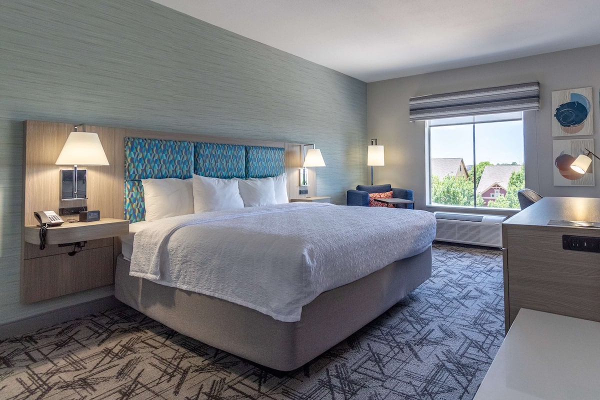 A hotel room with a large bed, two bedside tables with lamps, a desk, and a window with a view of buildings.