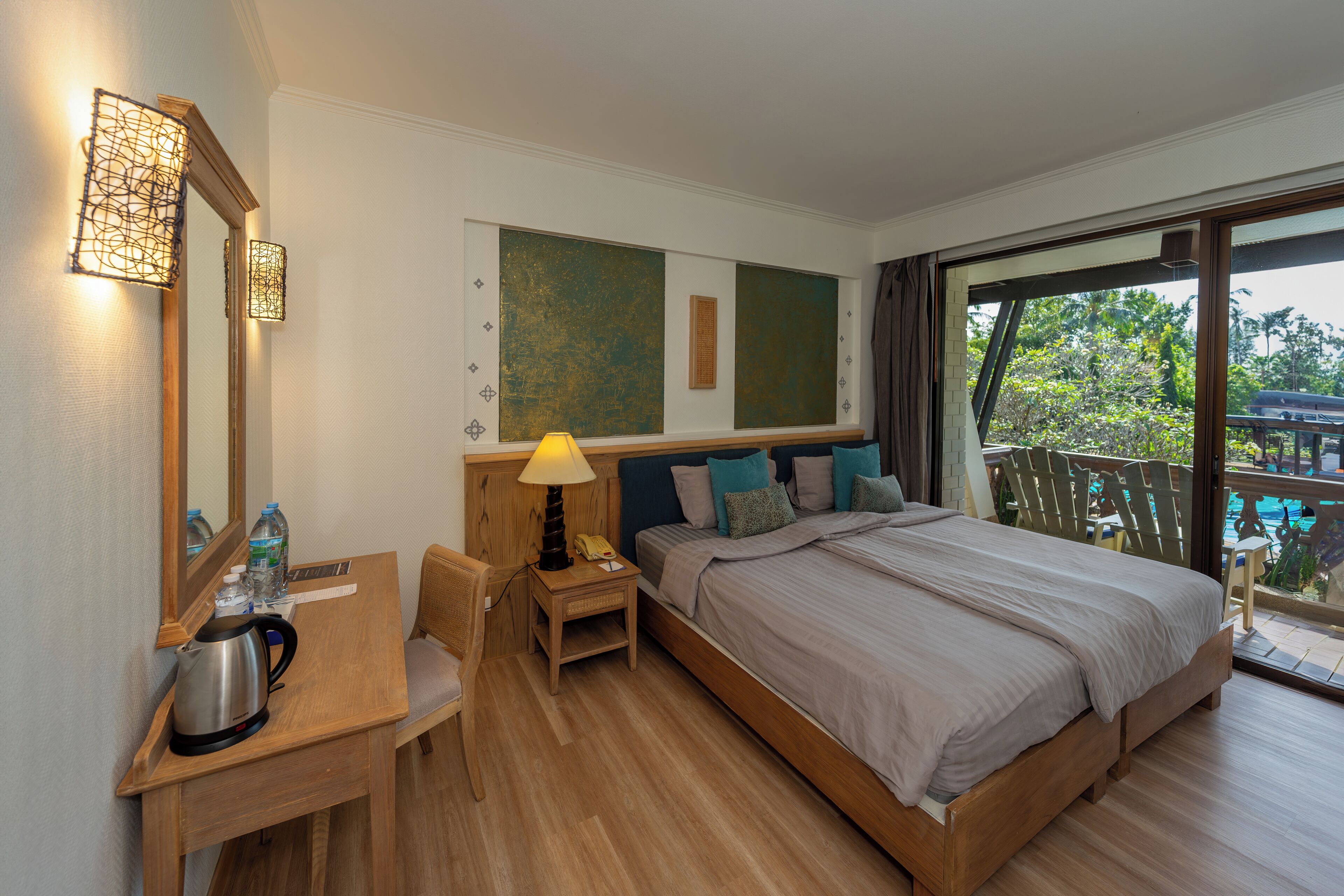 A hotel room with a bed, a desk, a chair, a lamp, a kettle, and a large window with a view of greenery.