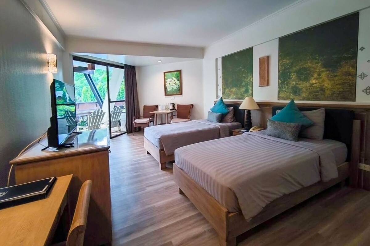 A hotel room with two beds, a desk with a computer, a chair, a small table, and a balcony with chairs.