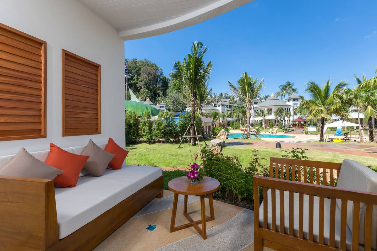 A modern outdoor seating area with a sofa and chairs, a small table with a vase, and a view of a pool and palm trees.