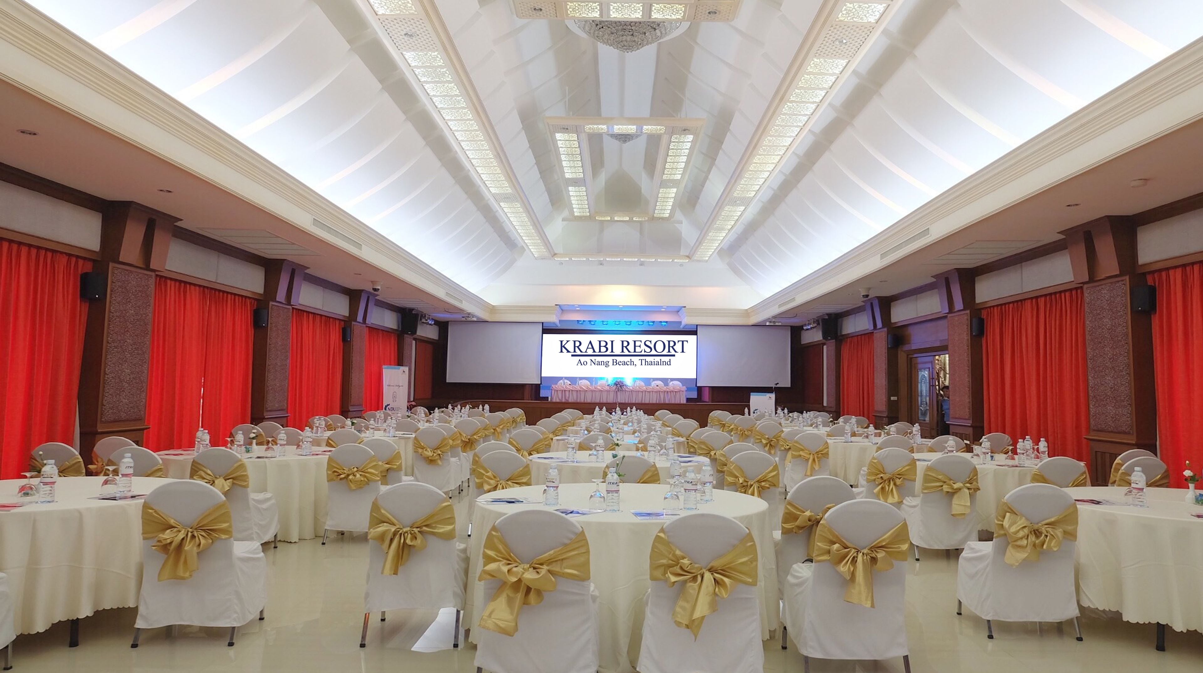 Un salón de banquetes con mesas redondas, manteles blancos y sillas adornadas con cintas doradas. Una pantalla grande muestra el texto: KRABI RESORT Ao Nang Beach, Tailandia.