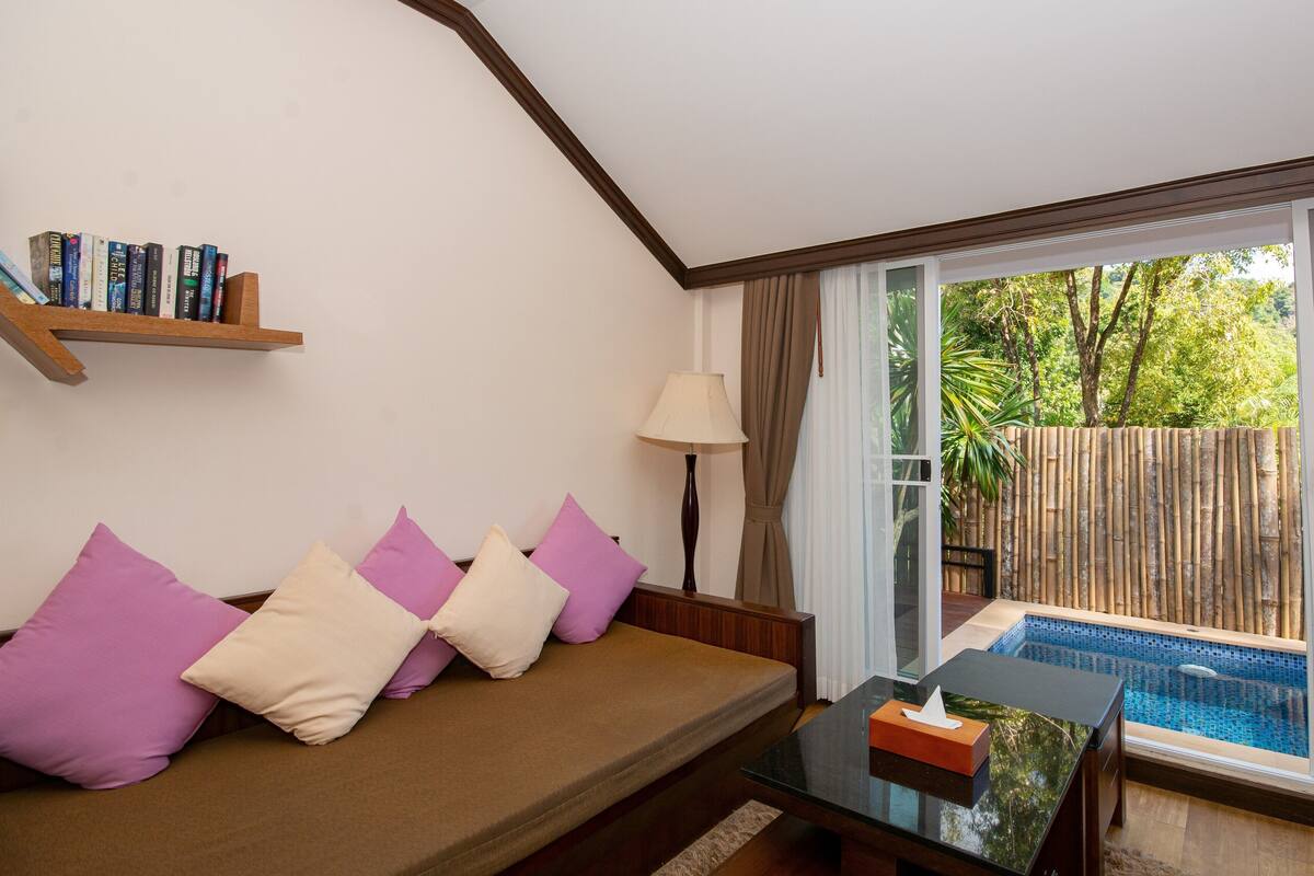 A cozy room with a sofa, a small table, a lamp, and a view of a swimming pool through large sliding doors.