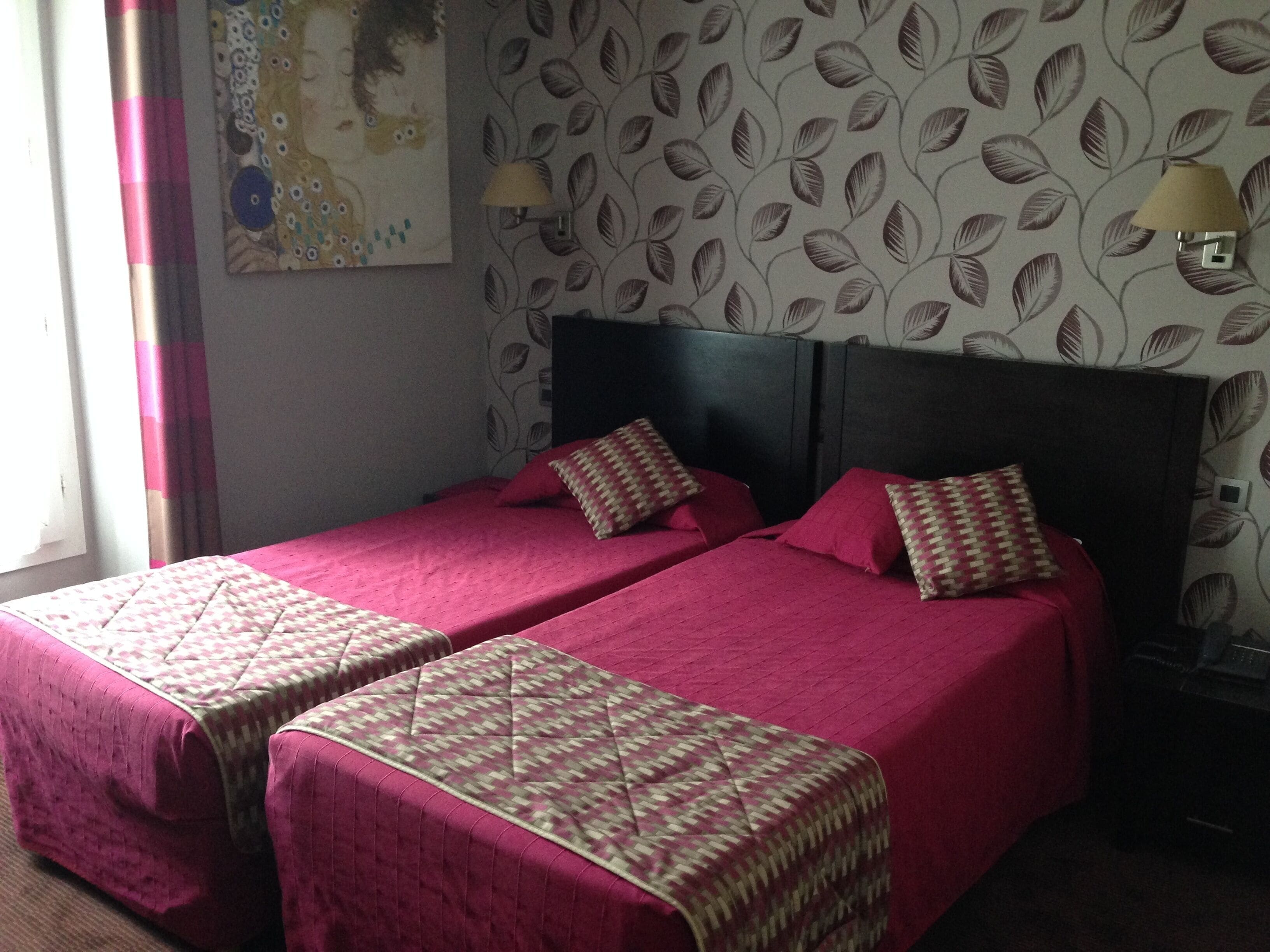 Une chambre d’hôtel avec deux lits, chacun recouvert d’une couverture rouge et agrémenté d’oreillers assortis. Un mur est décoré d’un motif de feuilles et un tableau est accroché au-dessus des lits.