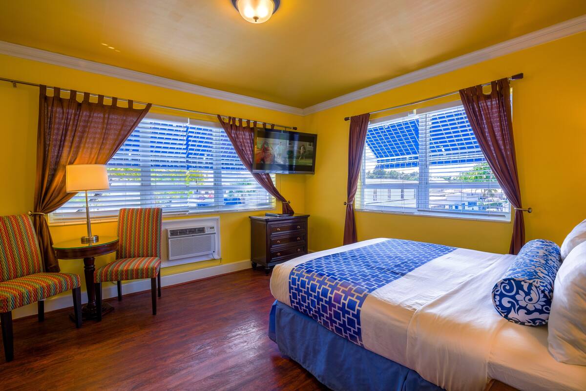 A hotel room with a bed, two chairs, a table, a TV, and a window with curtains.
