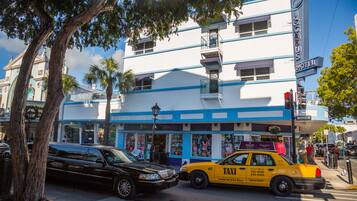A two-story building with a blue and white facade, a visible hotel sign, and a street corner with a taxi and a black car.