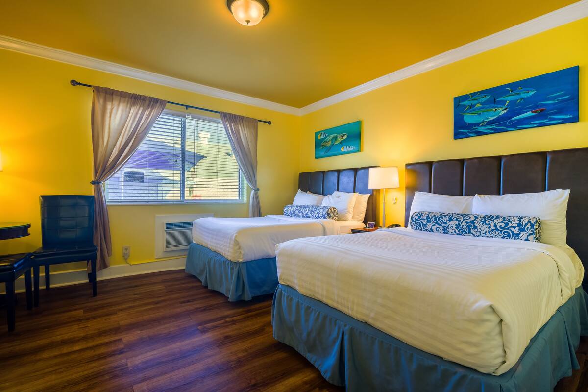 A hotel room with two beds, a desk, and a chair. There is a window with curtains, a painting on the wall, and a ceiling light.
