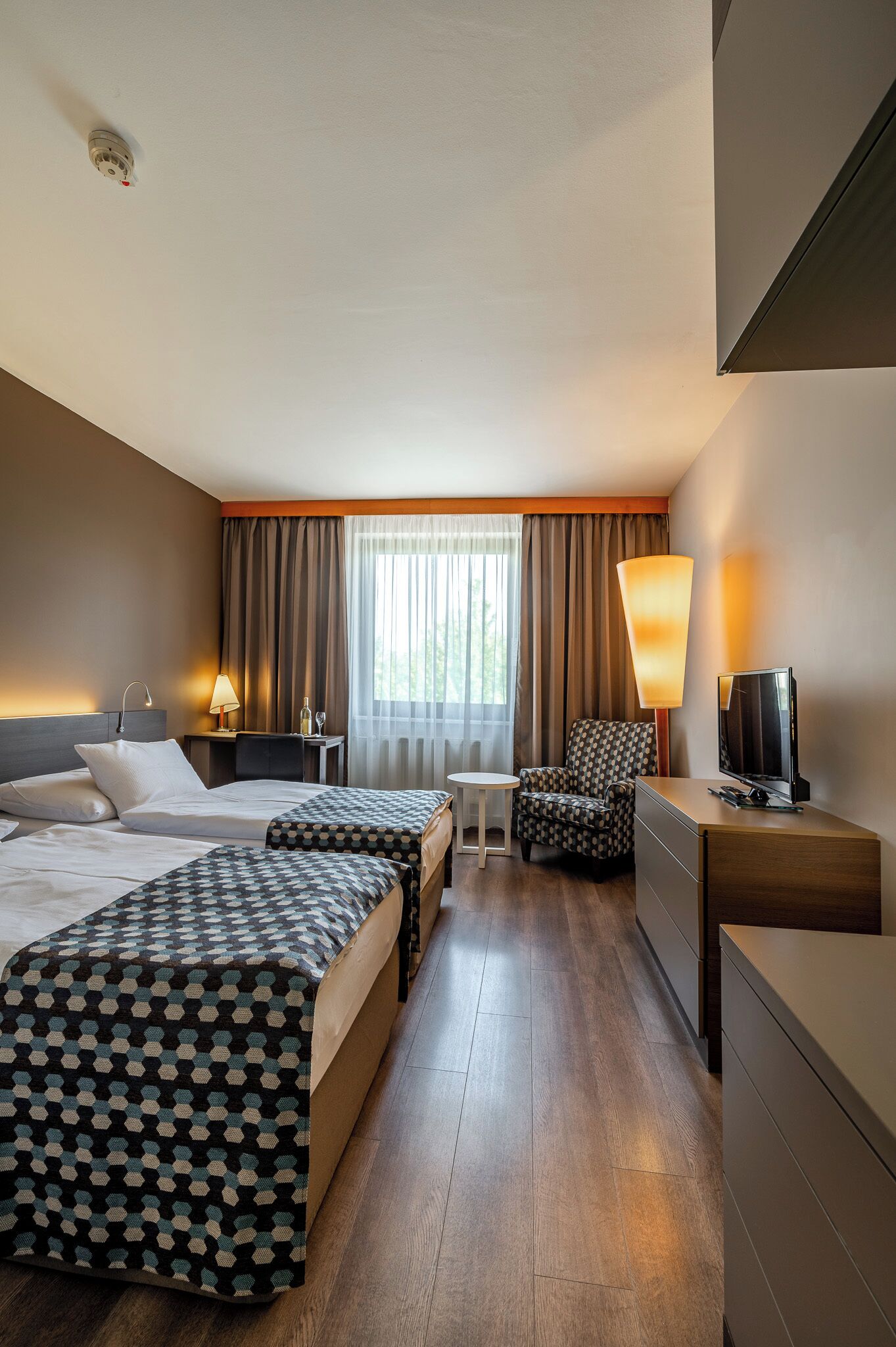 A hotel room with a bed, a desk, a chair, a TV, and a window with curtains.