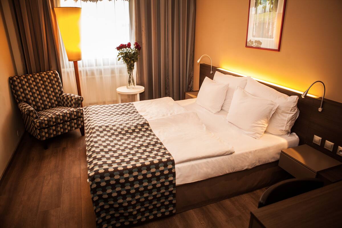 A hotel room with a large bed, a chair, a small table with a vase of flowers, and a nightstand with a lamp.