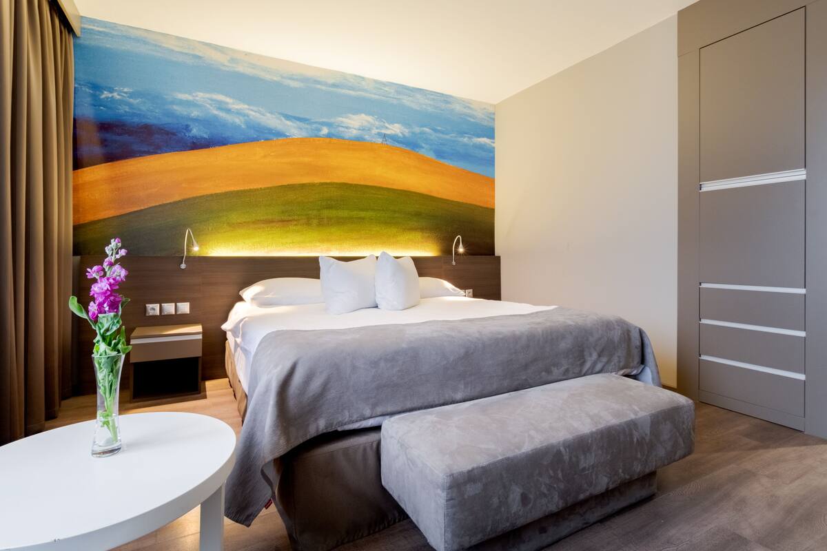 A modern hotel room with a large bed, a bedside table, a small table with a vase of flowers, and a wardrobe.