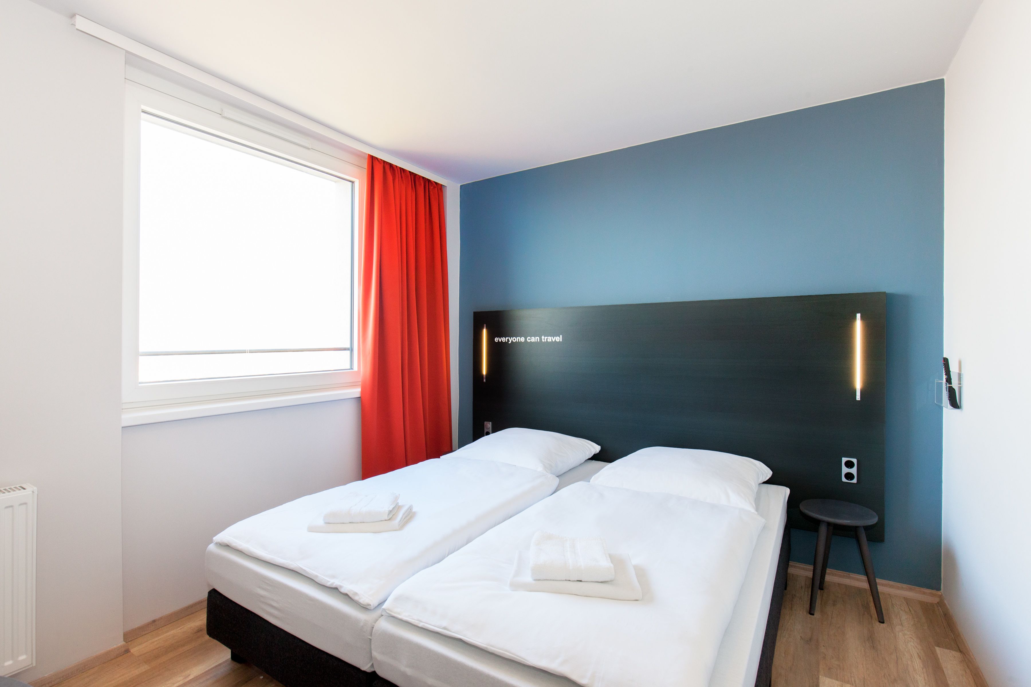 Une chambre d’hôtel avec deux lits, une fenêtre avec des rideaux rouges, un lampadaire fixé au mur et un petit tabouret.