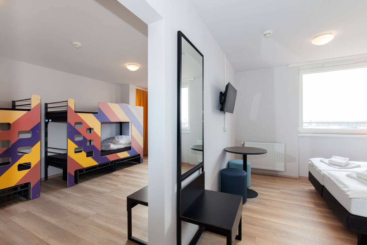 A modern hotel room with bunk beds, a small desk, a chair, a TV, and a large window.