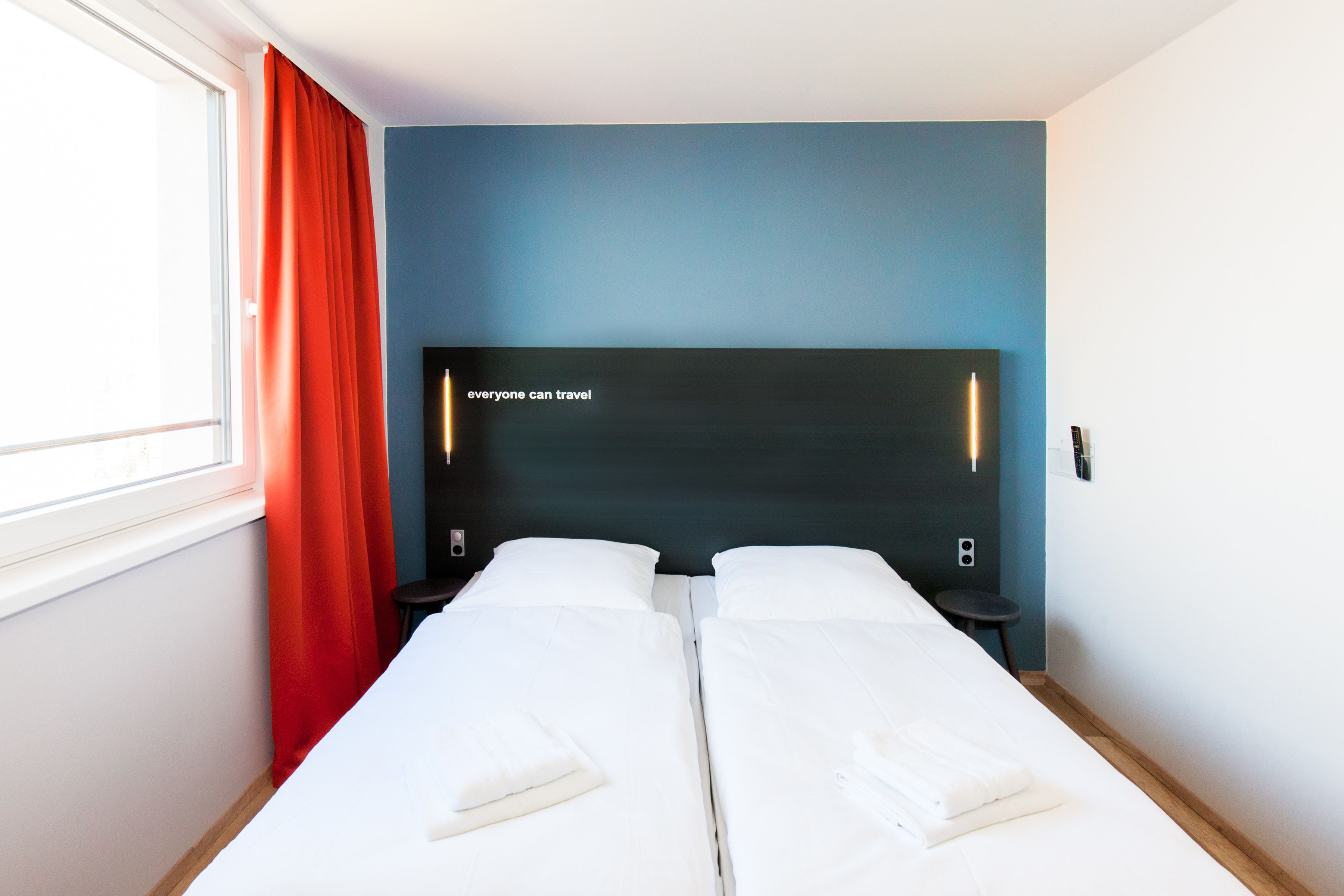 A double bed room with a dark headboard, a window with red curtains, and a wall-mounted light.