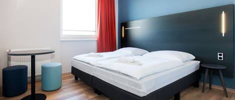 A modern hotel room with a bed, two side tables, a radiator, and a window with red curtains.