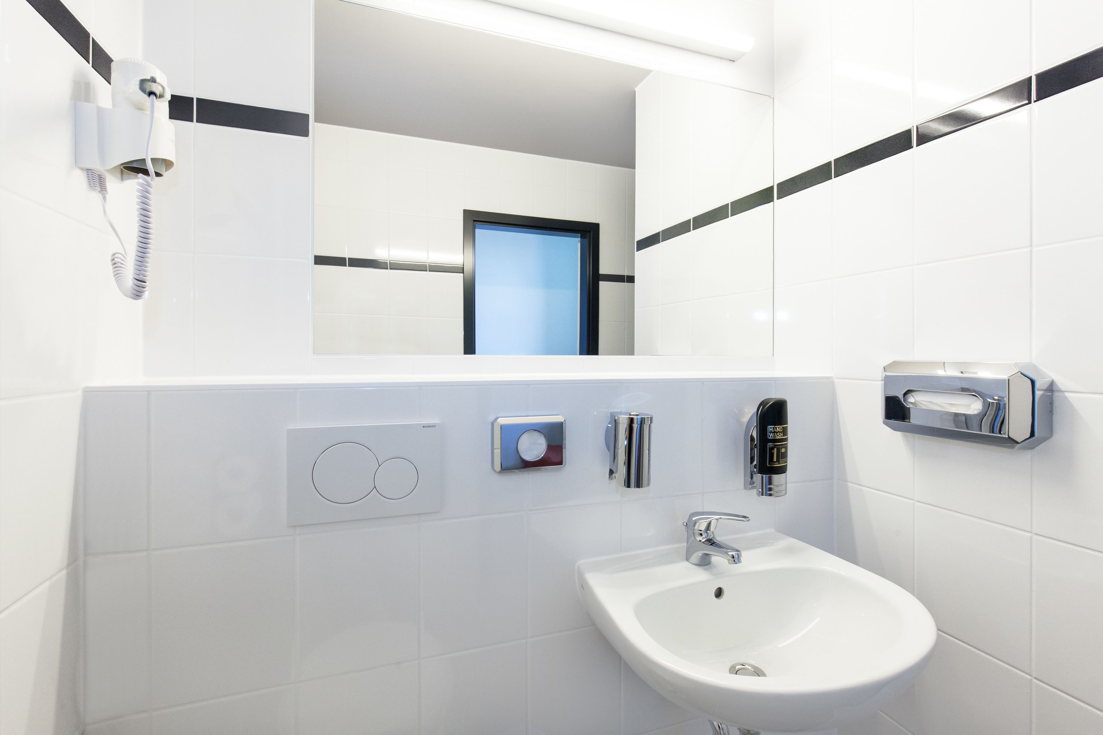 Ein modernes Badezimmer mit einem weißen Waschbecken, einem Spiegel, einem Handtuchspender und einem an der Wand montierten Föhn.