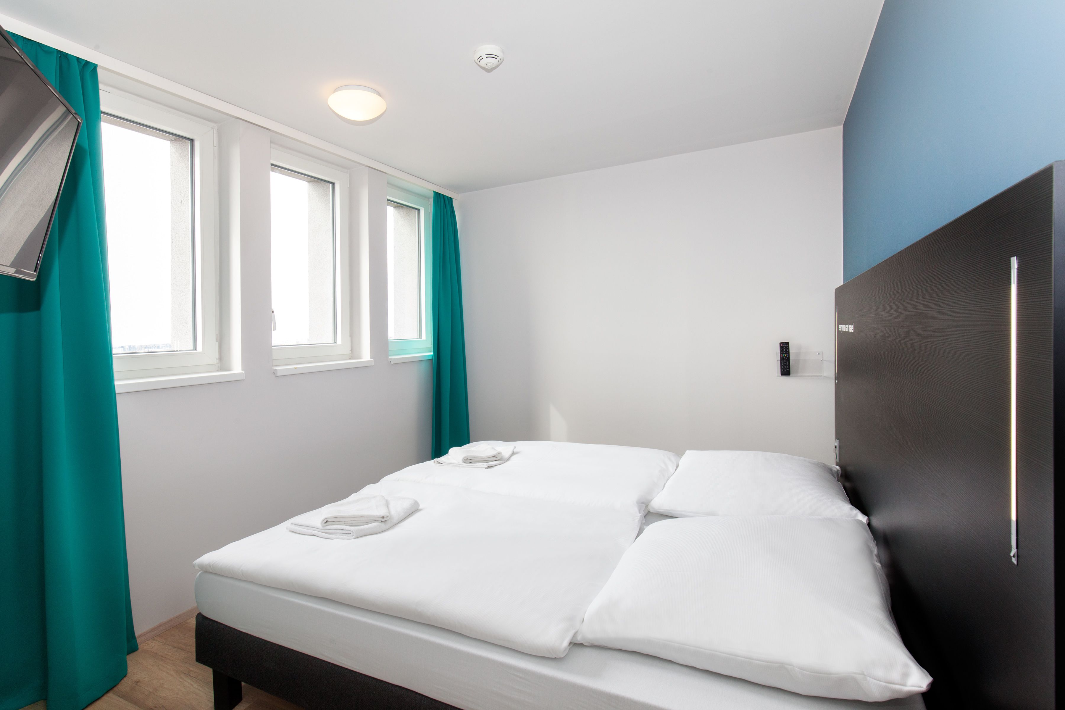 Una cama bien tendida con sábanas blancas, un cabecero oscuro y una ventana con cortinas azul verdosas.