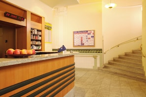 Zona de recepción de un hotel con un mostrador de madera, un reloj marcando el 22 de junio, 17:35, un tablón de anuncios con un mapa y una escalera.
