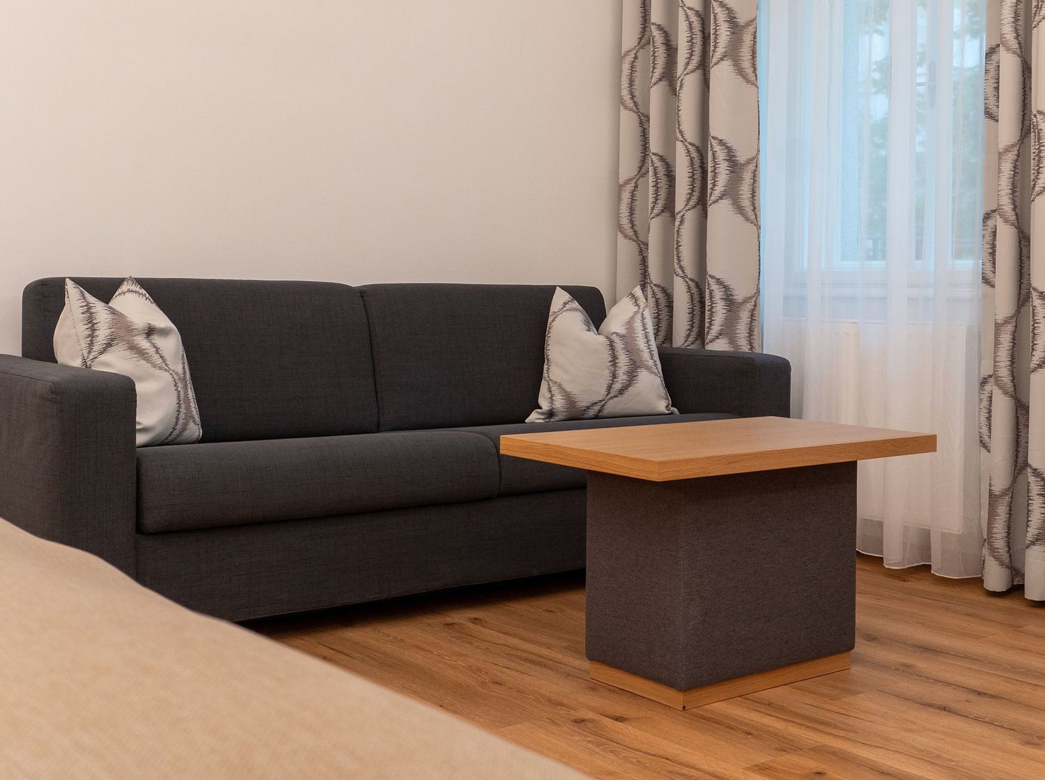Ein modernes Wohnzimmer mit einem grauen Sofa, zwei passende Kissen, einem Holztisch und transparenten Vorhängen mit Blättermotiv.