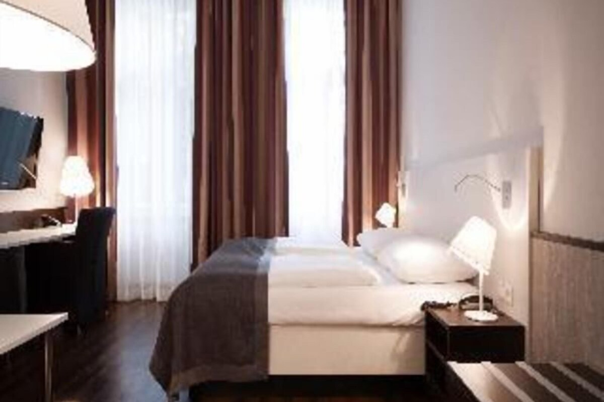 Quarto de hotel com cama, mesa de cabeceira, luminária, escrivaninha, cadeira e cortinas.