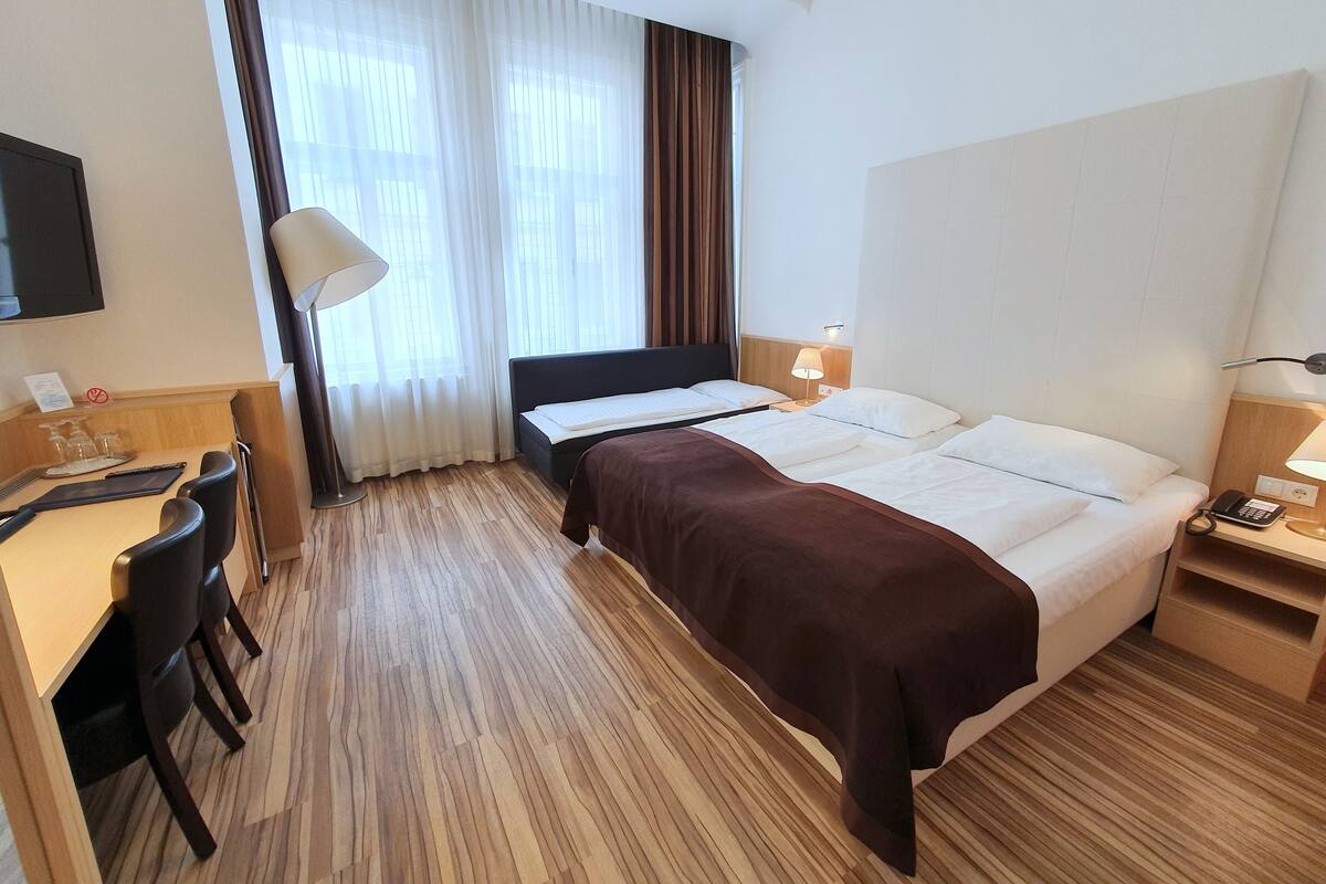 A hotel room with a bed, bedside table, lamp, desk, chair, and a TV on the wall.