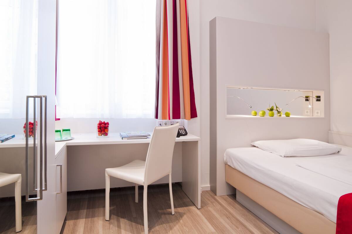 A modern hotel room with a bed, a desk, and a chair. There is a window with curtains, a mirror with a shelf, and a small table with a vase of flowers.