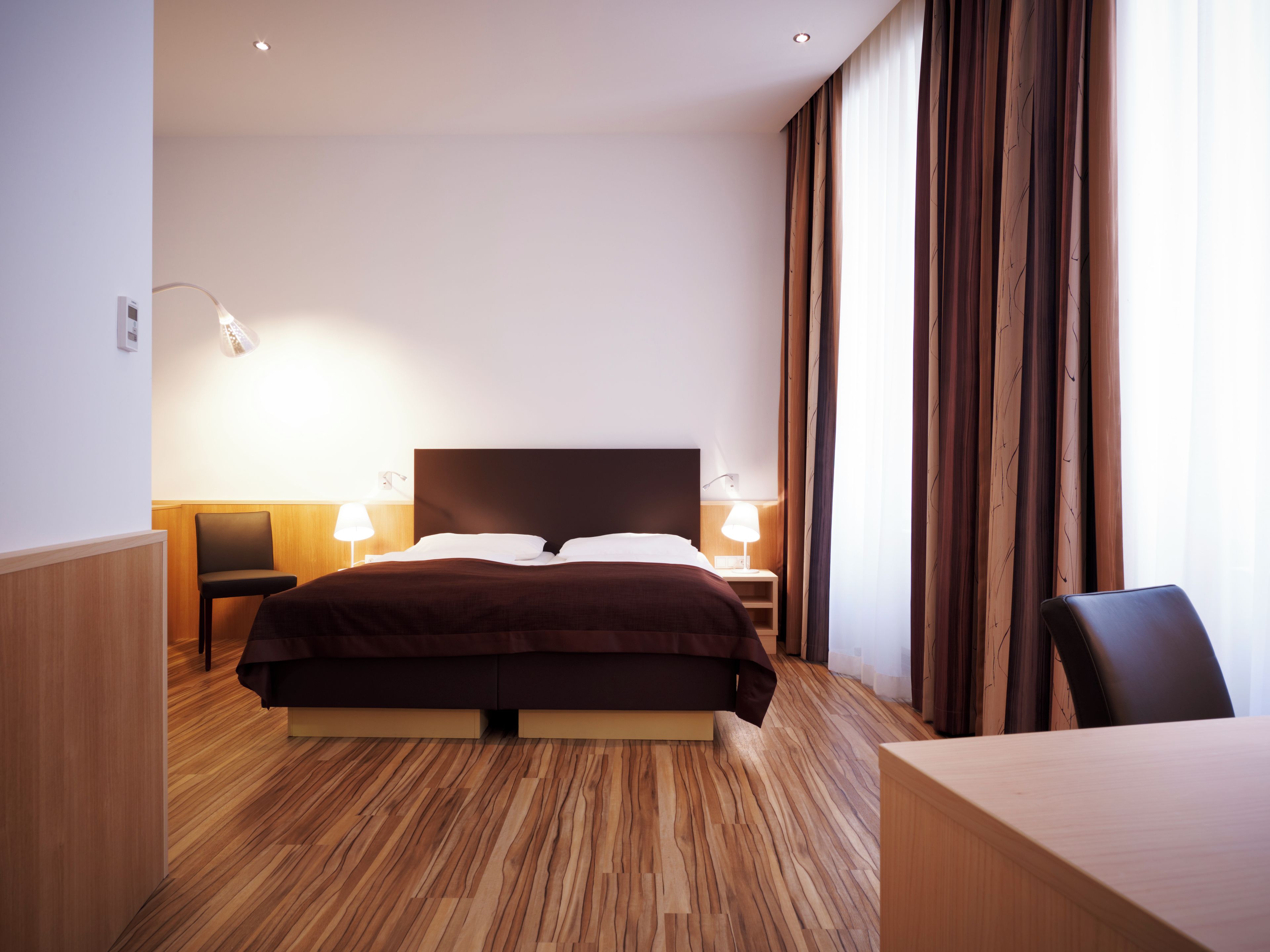 A hotel room with a large bed, two bedside tables with lamps, a wooden floor, and a view through sheer curtains.