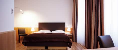 A hotel room with a large bed, two bedside tables with lamps, a wooden floor, and a view through sheer curtains.