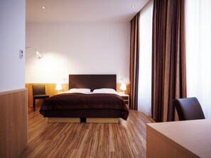 Hotellihuone, jossa on suuri sänky, kaksi yöpöytää valaisimineen, puulattia ja näkymä ohuiden verhojen läpi.