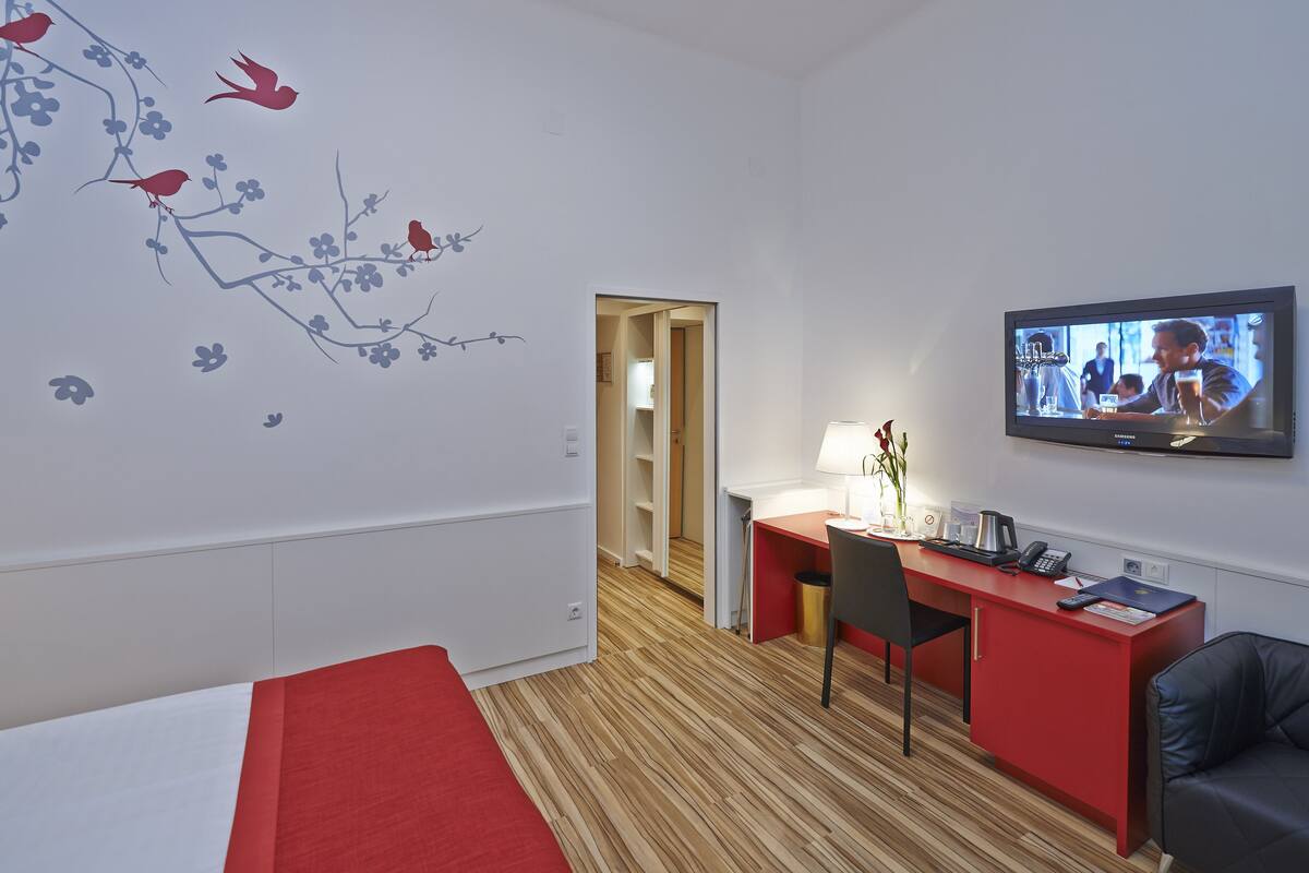 A hotel room with a bed, a desk, a chair, a TV, and a wall with floral decorations.