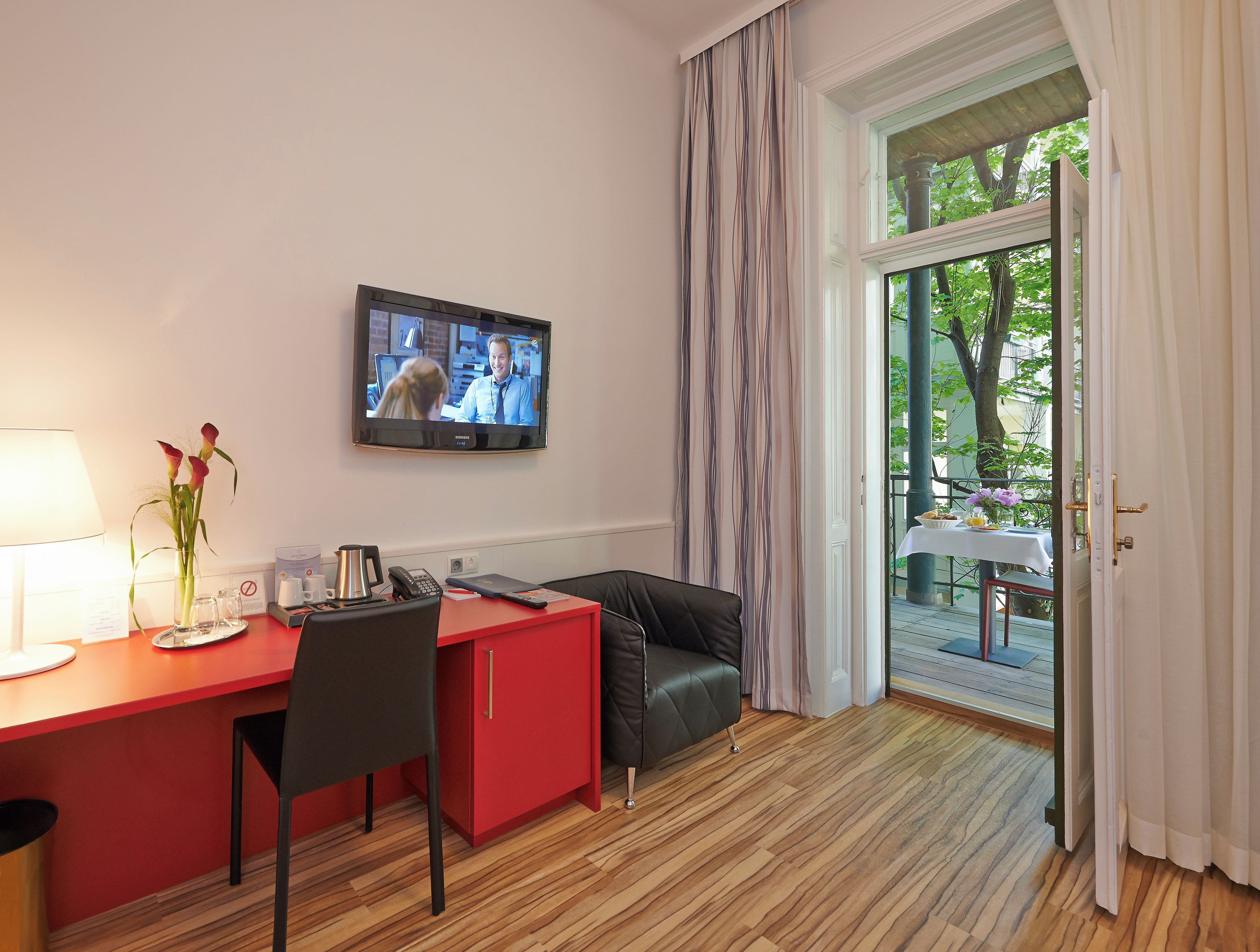 Een hotelkamer met een houten vloer, een rode bureau met stoel, een aan de muur bevestigde televisie en uitzicht op de omgeving door een groot raam.