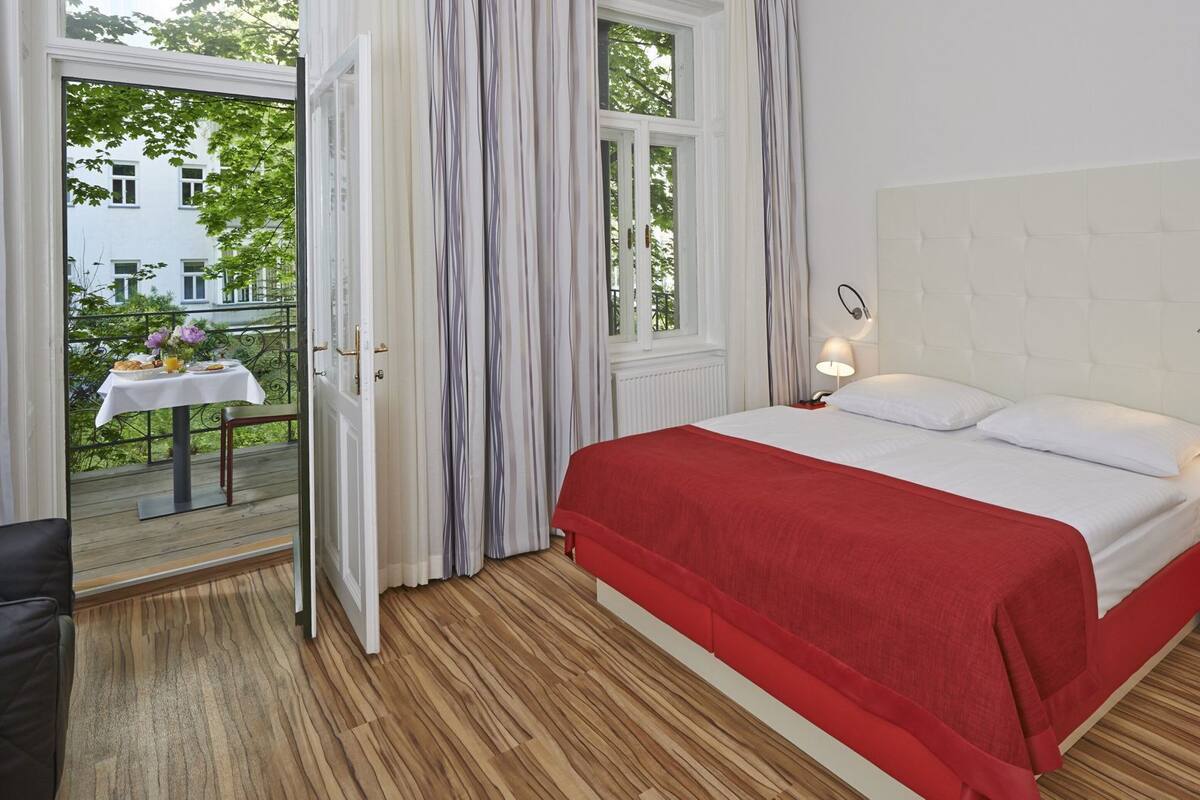 A bedroom with a bed, two bedside lamps, a window with curtains, a view of the outside, and a small table with a flower arrangement.