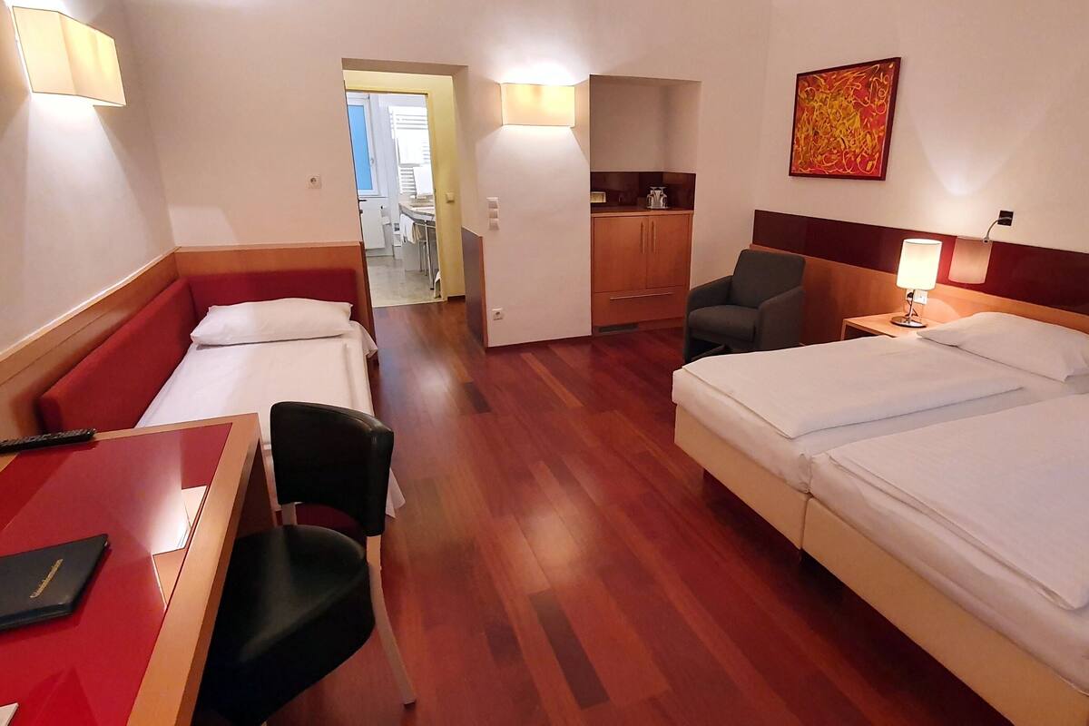 A hotel room with a large bed, a desk with a chair, a small table, and a kitchenette area.