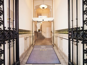 Entrada de hotel con una escalera de mármol, barandillas de hierro forjado negro y una alfombra azul con el nombre del hotel.
