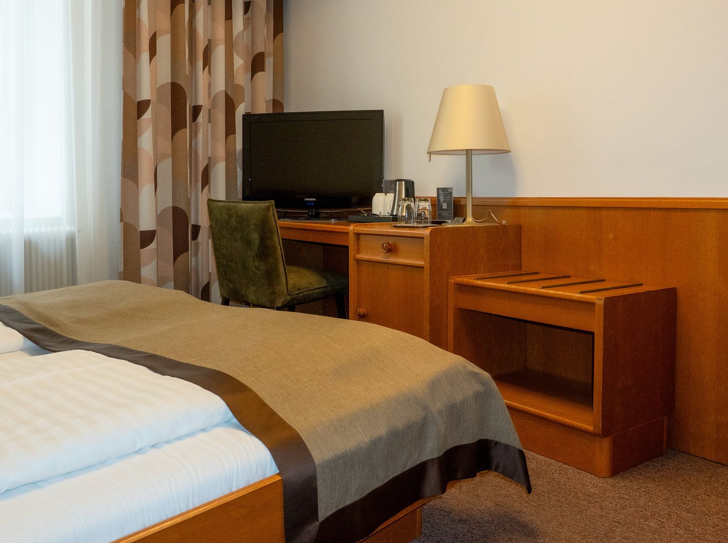 Ein Hotelzimmer mit einem Bett, einem Schreibtisch mit Fernseher, einem Sessel, einer Lampe und einem kleinen Regal.
